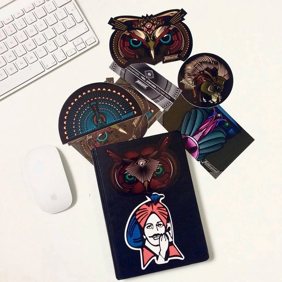 Assorted colorful sticker sheets featuring owl and animal designs, a black box with a sticker of a woman with glasses and red hair, a white Apple Magic Mouse, and part of a white keyboard on a white surface.