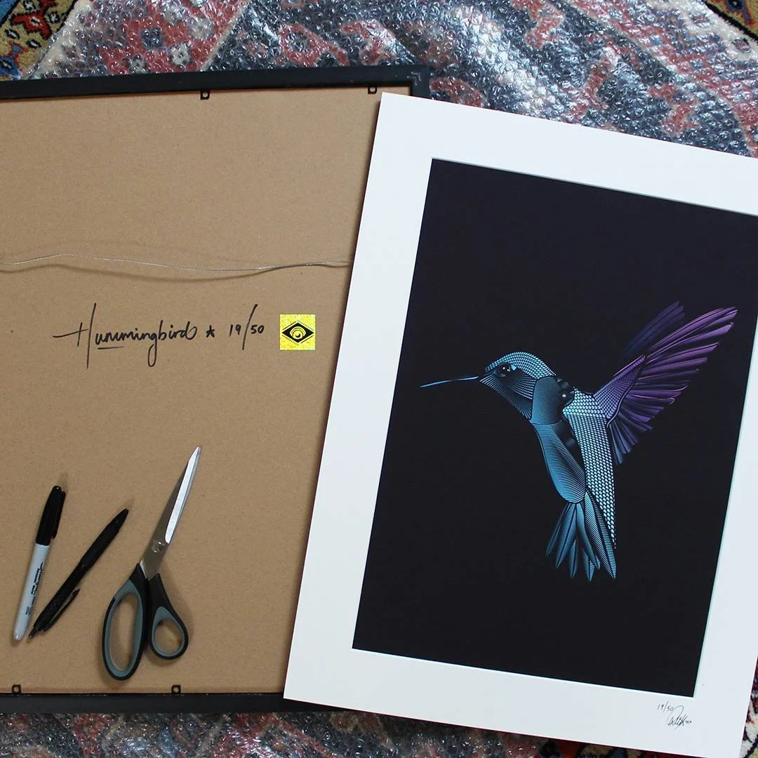 The image shows a large print of a digital illustration of a hummingbird with metallic, geometric features and purple wings, lying on a dark background. Next to it are crafting tools including scissors, a black marker, a white pen, and a wire, on a t
