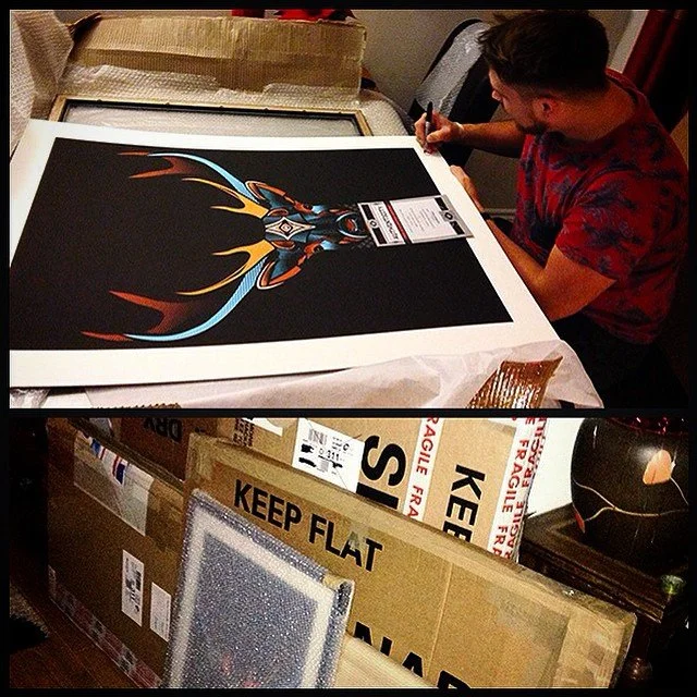 A person is signing a large, colorful artwork featuring a stylized, futuristic figure with flames or energy emanating from it, on a table covered with white paper. Surrounding the table are cardboard boxes labeled "Keep Flat" and "Fragile," indicatin