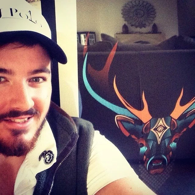 A man with a beard and mustache smiling, wearing a white cap and a jacket, sitting next to a pillow with a colorful, stylized deer head design by Fieldinspired.