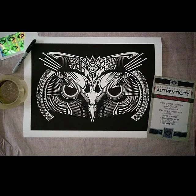 Black and white art of an owl with geometric patterns on white paper, next to a certificate of authenticity. There is also a tattoo needle and ink container on the table.