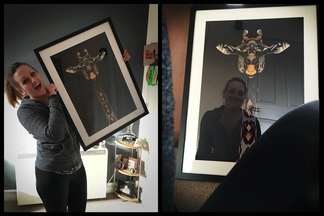 A woman holding a framed art piece with a stylized animal face in black, white, and orange. The woman is smiling in the left photo and reflected in the glass of the frame in the right photo.