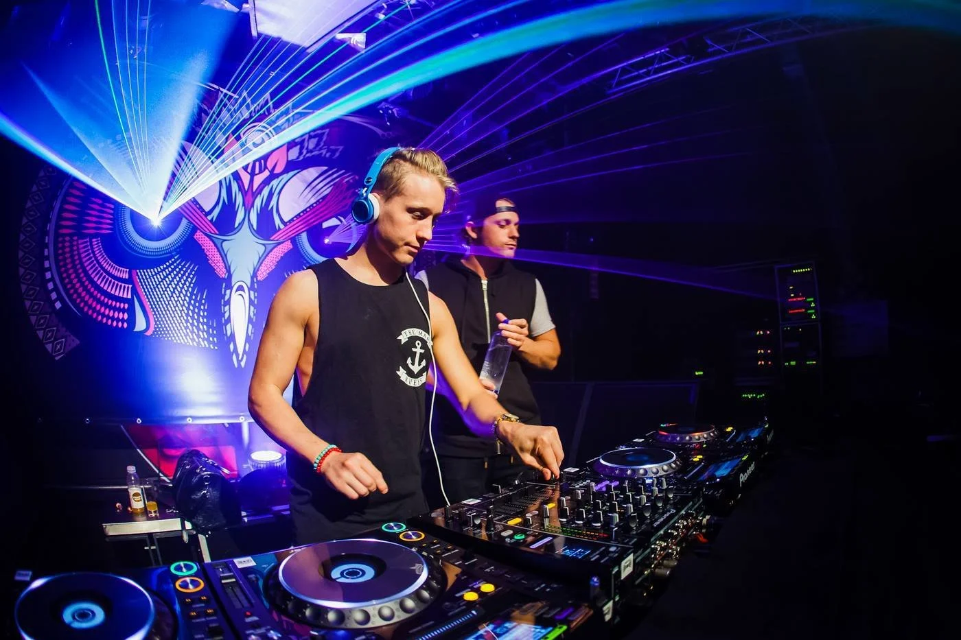 Two male DJs perform in a nightclub, with vibrant laser lights and a decorative backdrop behind them.