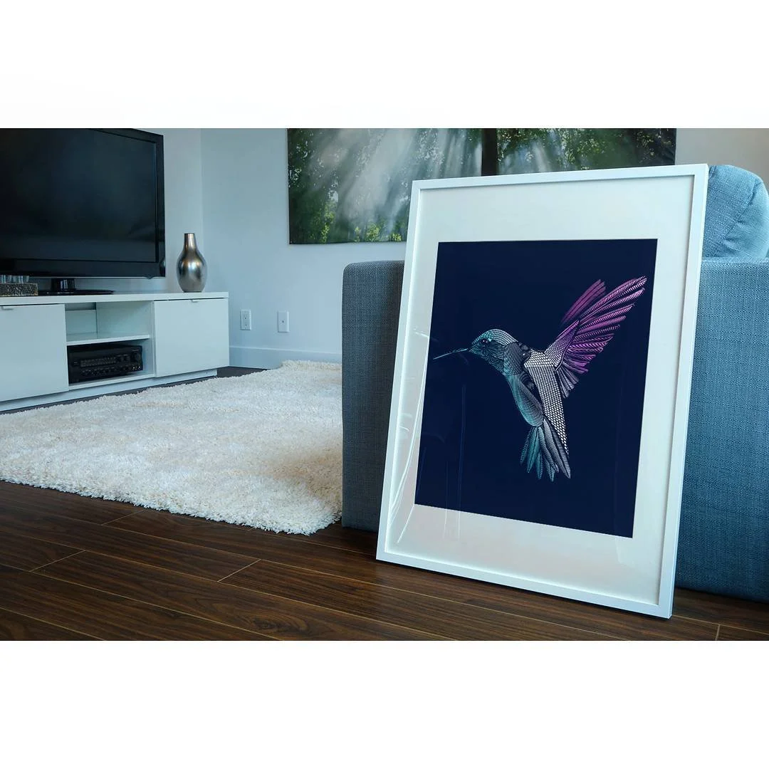 Framed artwork of a hummingbird resting against a blue background leaning against a gray sofa in a living room with a white media console, a large flat-screen TV, and a vase, with a window showing trees and sunlight.