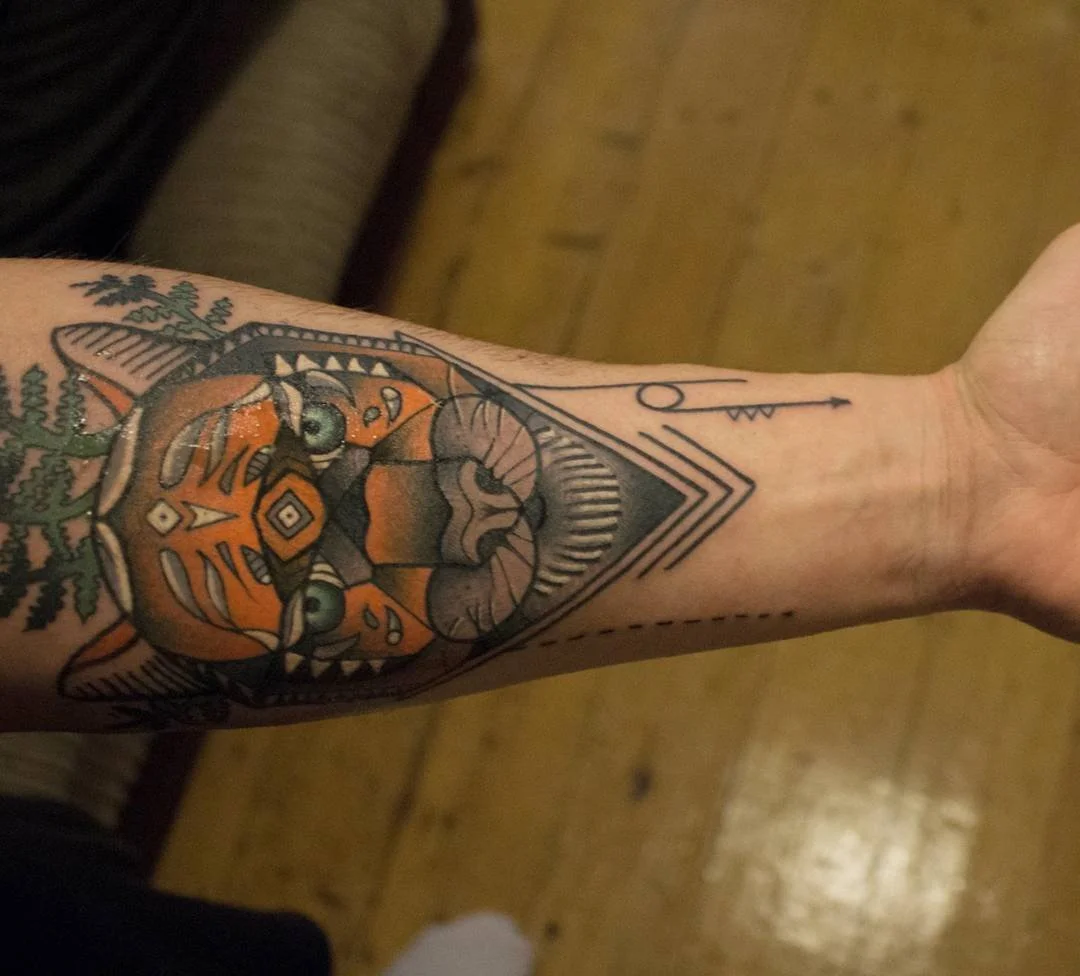 Tattoo of a cat's face with tribal design elements, with tree branches to the left and geometric shapes around it.