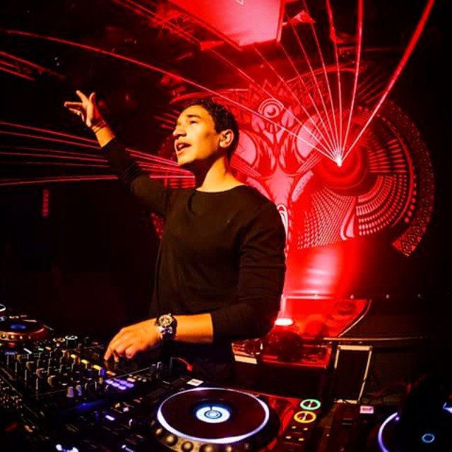 A DJ performs under red laser lights and digital graphics in a club or event space.