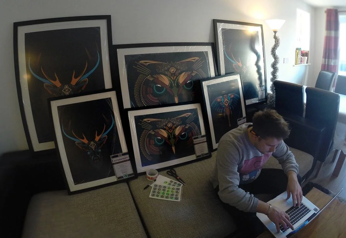 A person sitting on a couch using a laptop, with framed artwork of colorful deer heads leaning against the wall behind them. The room has a standing lamp, books on a shelf, a table with scissors and stickers, and a window with curtains.