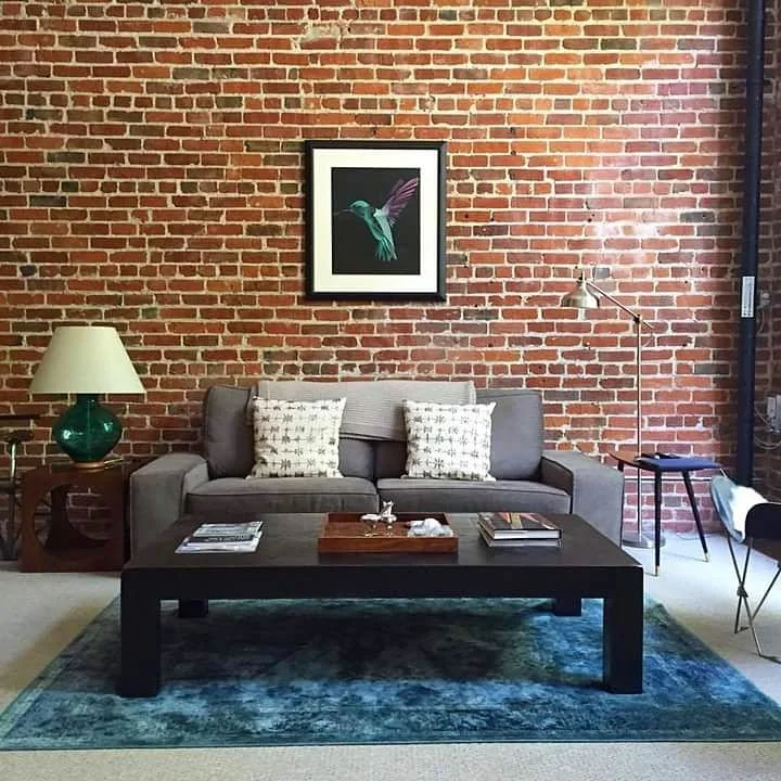 Photo sent in by a customer of their living room with a brick wall background, gray sofa with three pillows, a black coffee table with books and a decorative tray, a framed hummingbird by fieldinspired print on the brick wall.