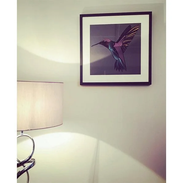 A framed artwork of a colorful hummingbird hangs on a light-colored wall in a room. A table lamp with a beige shade is partially visible in the foreground, casting light onto the wall.