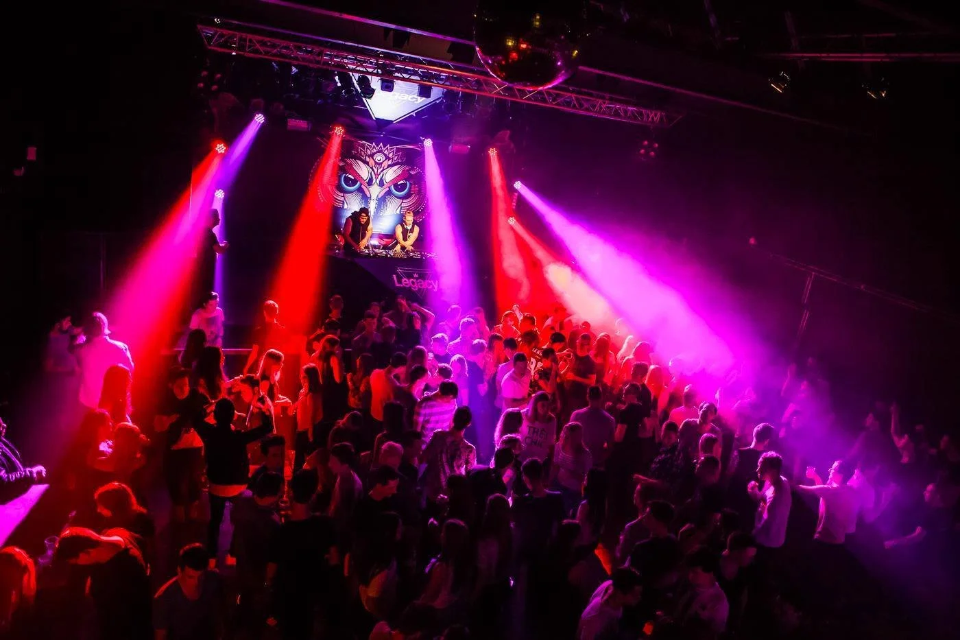 Crowd of people dancing at a nightclub with vibrant purple and red party lights, DJ booth with two DJs, and a large owl face graphic on a screen in the background.