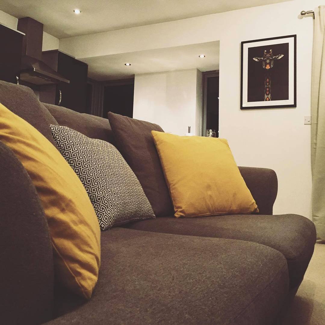 A cozy living room with a dark gray sofa adorned with three throw pillows, two yellow and one patterned black and white. In the background, there's a white wall with a framed artwork of a giraffe.