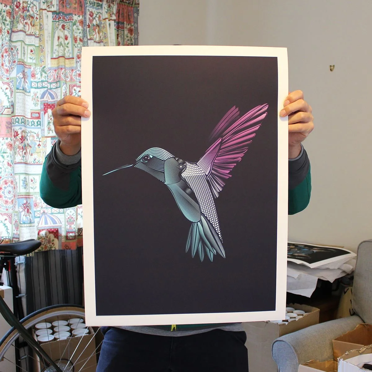 Person holding an art print of a stylized hummingbird with pink and blue hues on a dark background.