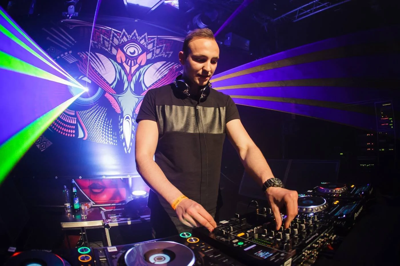 A DJ performing at a nightclub with colorful laser lights and a vibrant digital backdrop of a stylized face with an eye in the center.