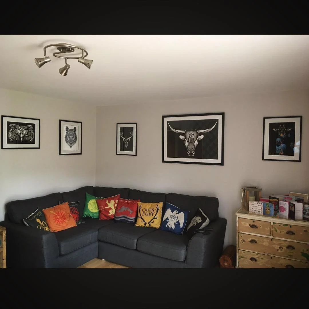A living room with a dark gray sofa decorated with colorful Game of Thrones themed pillows, a wooden dresser with books and cards, wall art of various animal faces, and a ceiling light fixture.