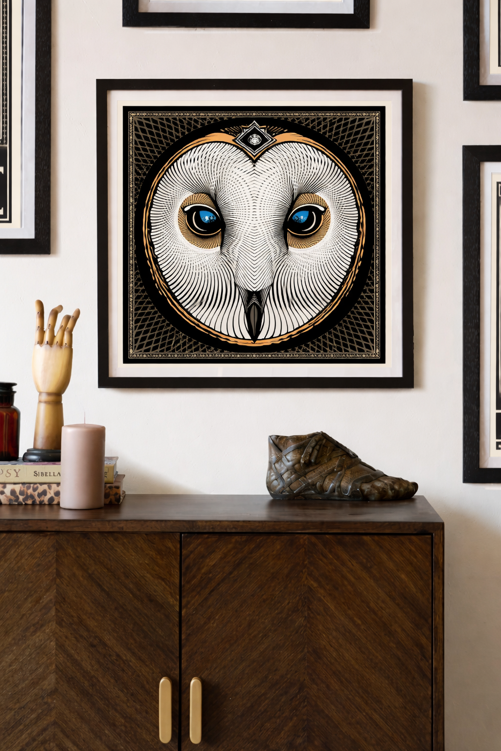 A framed black and white art piece resembling an owl's face with blue eyes, hanging on a white wall. Below the artwork is a wooden cabinet with a carved wooden shoe sculpture, a beige candle, and a few books on it.