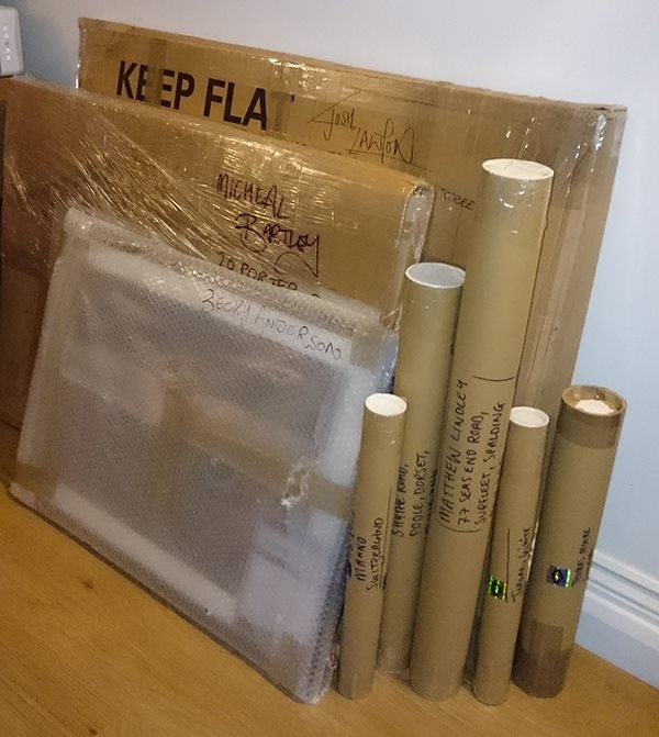 Multiple cardboard boxes and rolled-up posters or documents stacked against a wall and on a wooden floor, with handwritten labels on the boxes.