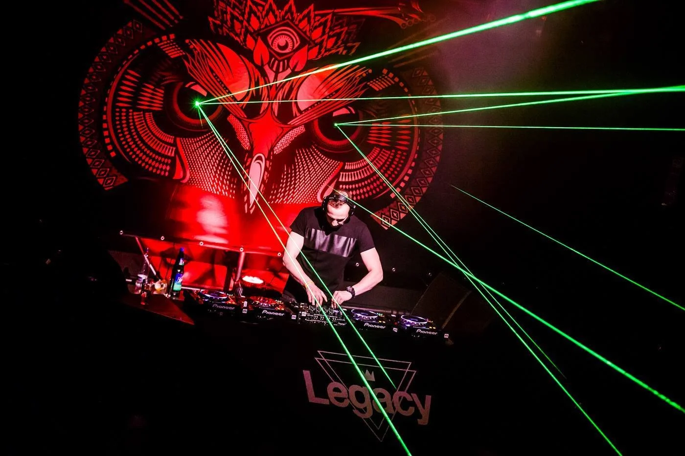 A DJ performing at a nightclub with green laser lights and a red artistic backdrop featuring an eye and tribal designs. The DJ is focused on mixing music behind Pioneer DJ equipment, with the word 'Legacy' visible on the front.