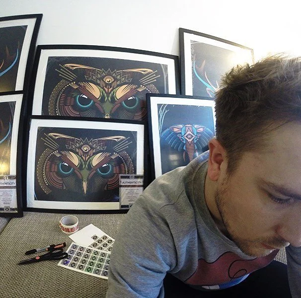 A young man with short, light brown hair and a beard, sitting in front of framed digital art prints of an owl and an elephant with vibrant, intricate designs. Art supplies and stickers are on the floor nearby.