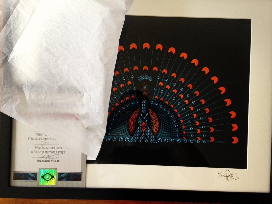 A framed artwork featuring a stylized peacock with elaborate tail feathers in red, blue, and black against a black background. The frame also contains a signed print, numbered 2/25, with a certificate of authenticity.