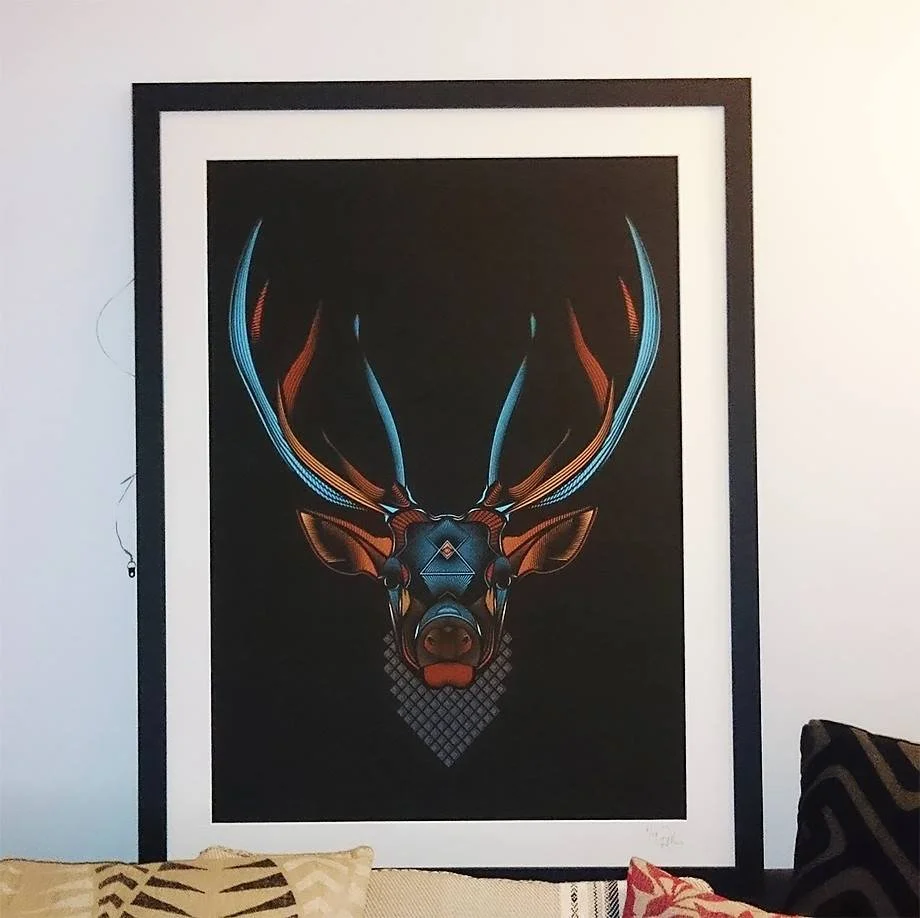 Framed digital artwork of a stylized deer head with antlers, featuring blue and orange geometric lines on a black background, hanging on a wall.