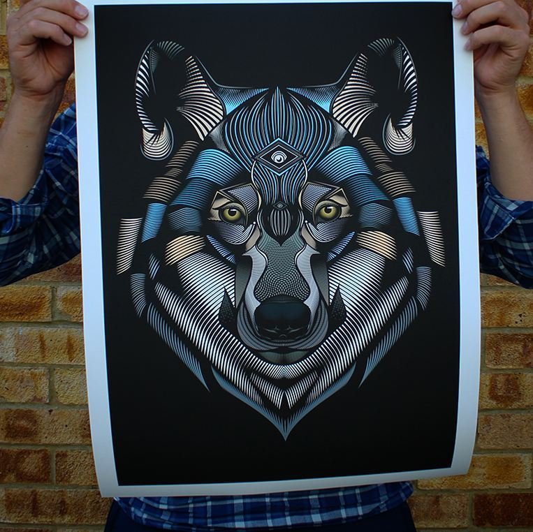 A person in a plaid shirt holding up a digital art print of a stylized wolf's face with glowing yellow eyes, intricate black, white, and blue line work, set against a black background.