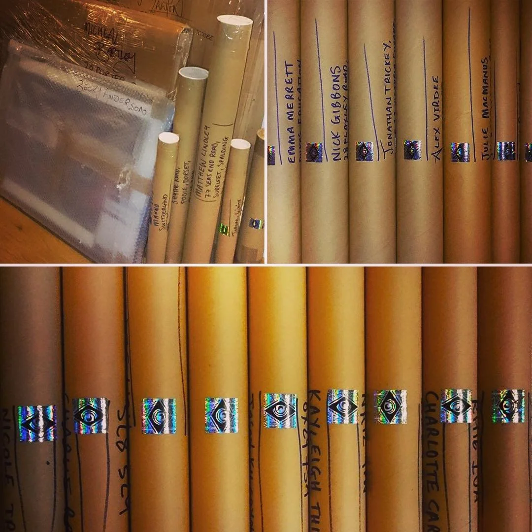 Several brown paper tubes with handwritten names and titles, stored upright, with labels and holographic stickers, likely used as scrolls or posters, on a wooden surface.