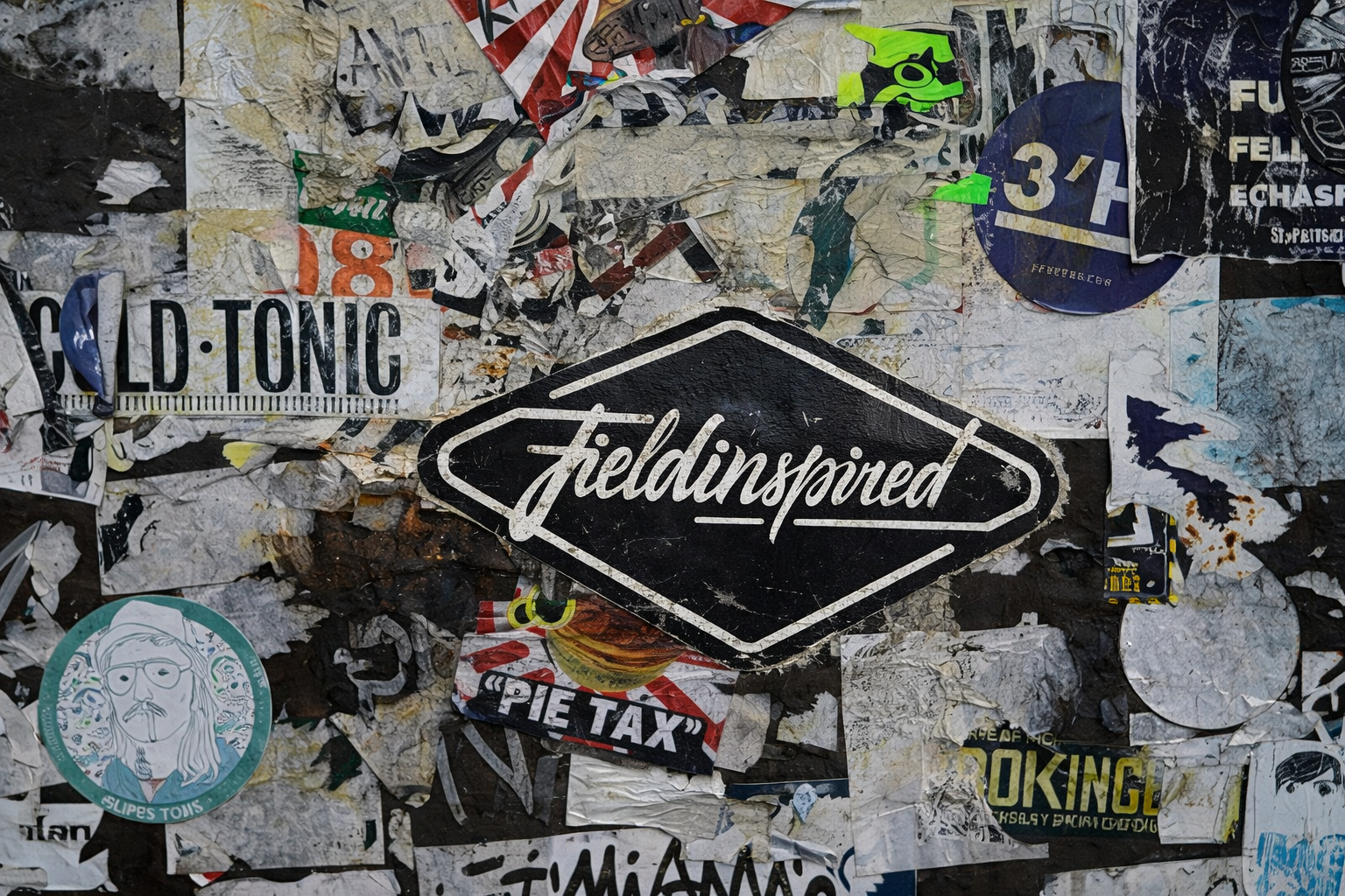 A collage of torn and layered stickers and posters on a surface, with a prominent black and white sticker that says 'Fieldinspired' in stylized script.