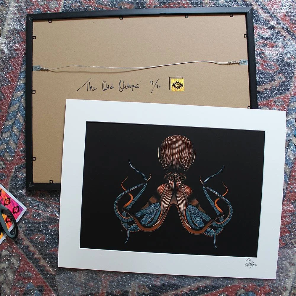 A framed, colorful illustration of an octopus against a black background, with the back of the artwork facing up and the octopus visible on the front. The back of the frame has a wiring setup for hanging and is signed with handwritten notes including