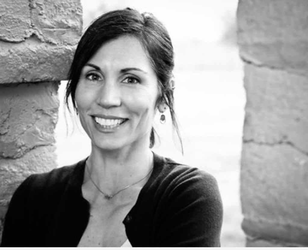 Black and white portrait of a woman smiling, with dark hair and earrings, standing between brick walls.