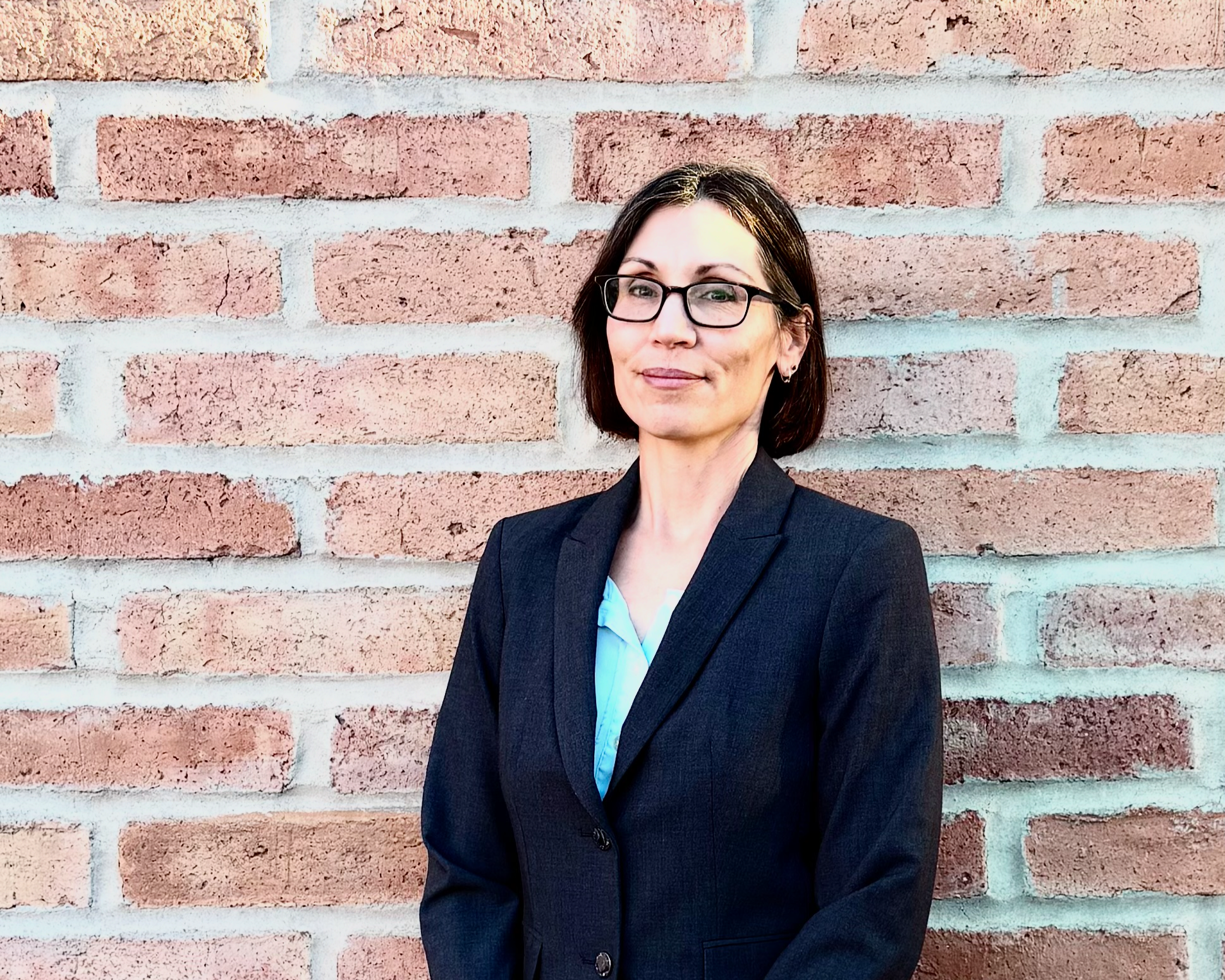 A woman with short dark hair, glasses, and a blazer standing against a brick wall.