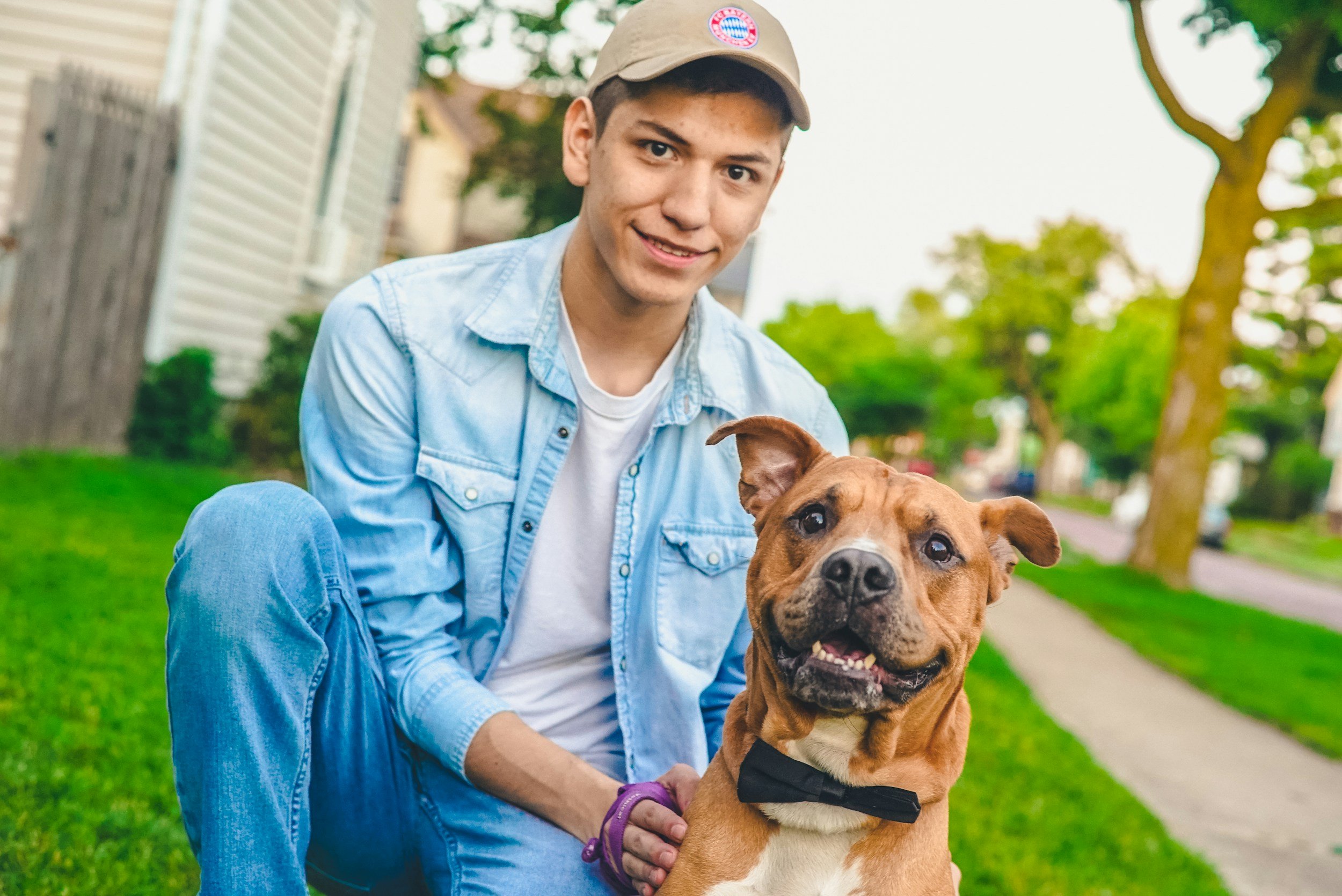 A young man with a light blue denim shirt and beige baseball cap sitting outdoors on grass with a brown dog wearing a black bow tie. The young man is smiling at the camera, and the background shows a suburban neighborhood with trees, houses, and a sidewalk.