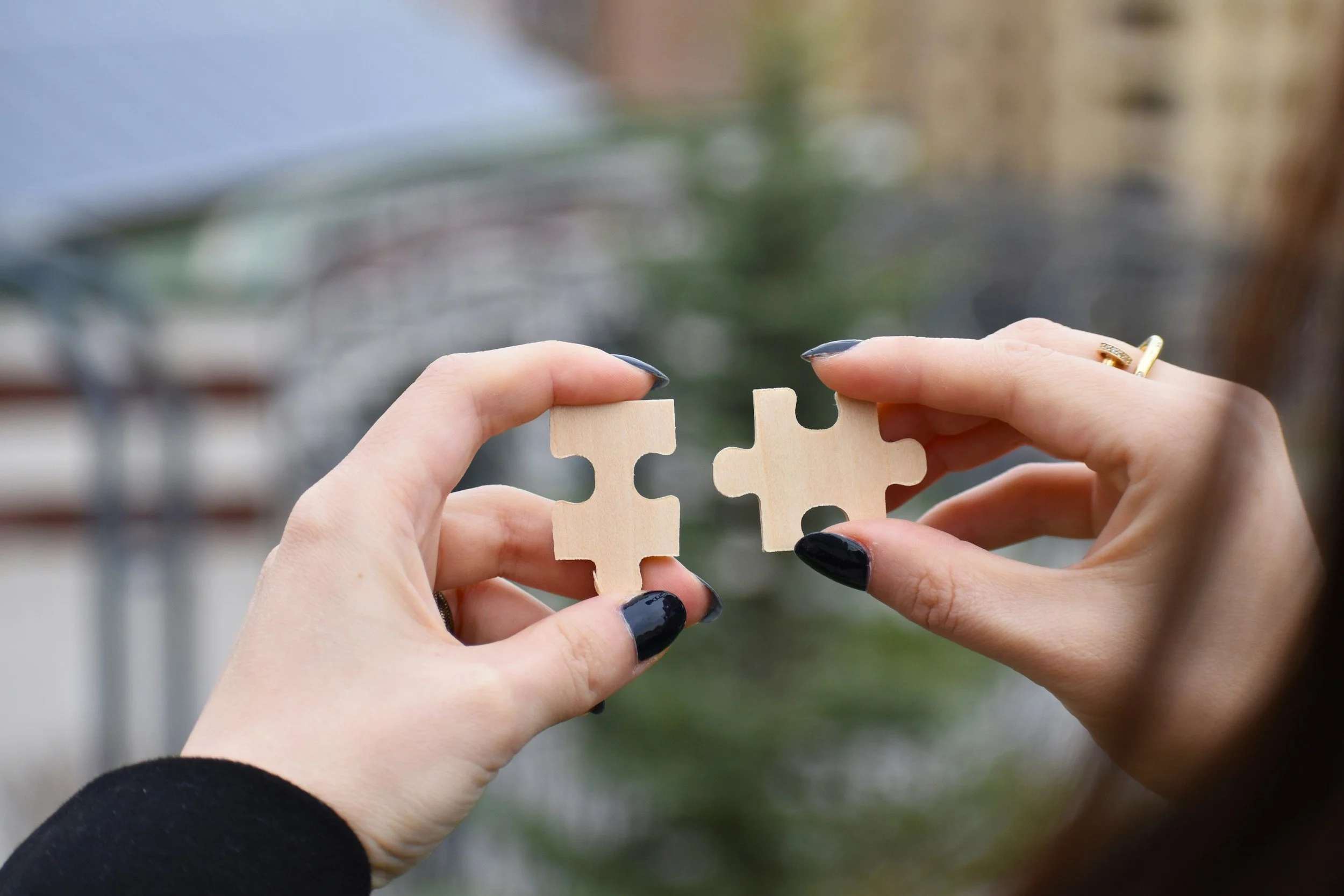 Two hands holding beige puzzle pieces, fitting them together against a blurred outdoor background.