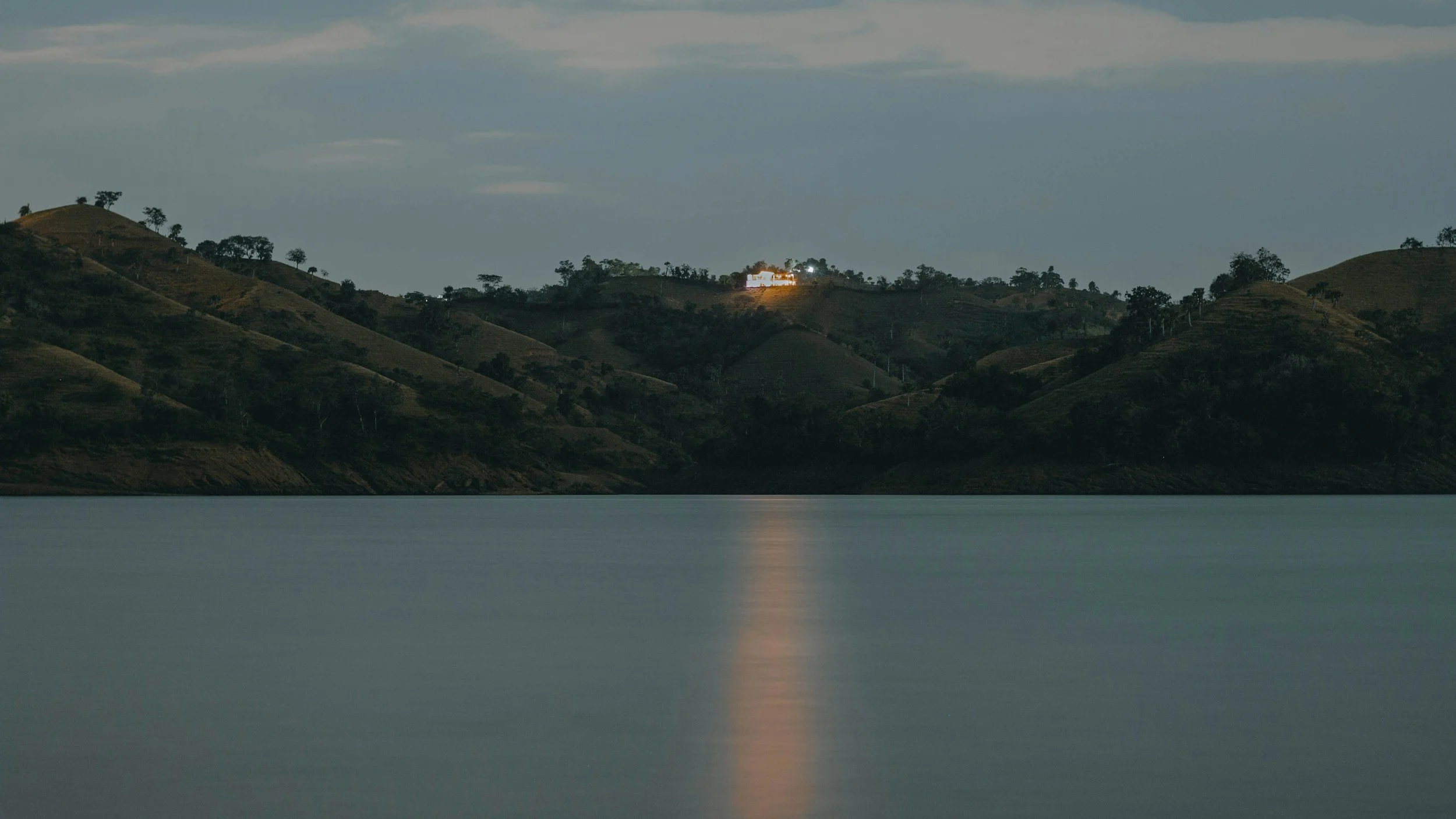 Nighttime view of a body of water with a hillside in the background,-lit building on top, and a reflection of the building's lights on the water.
