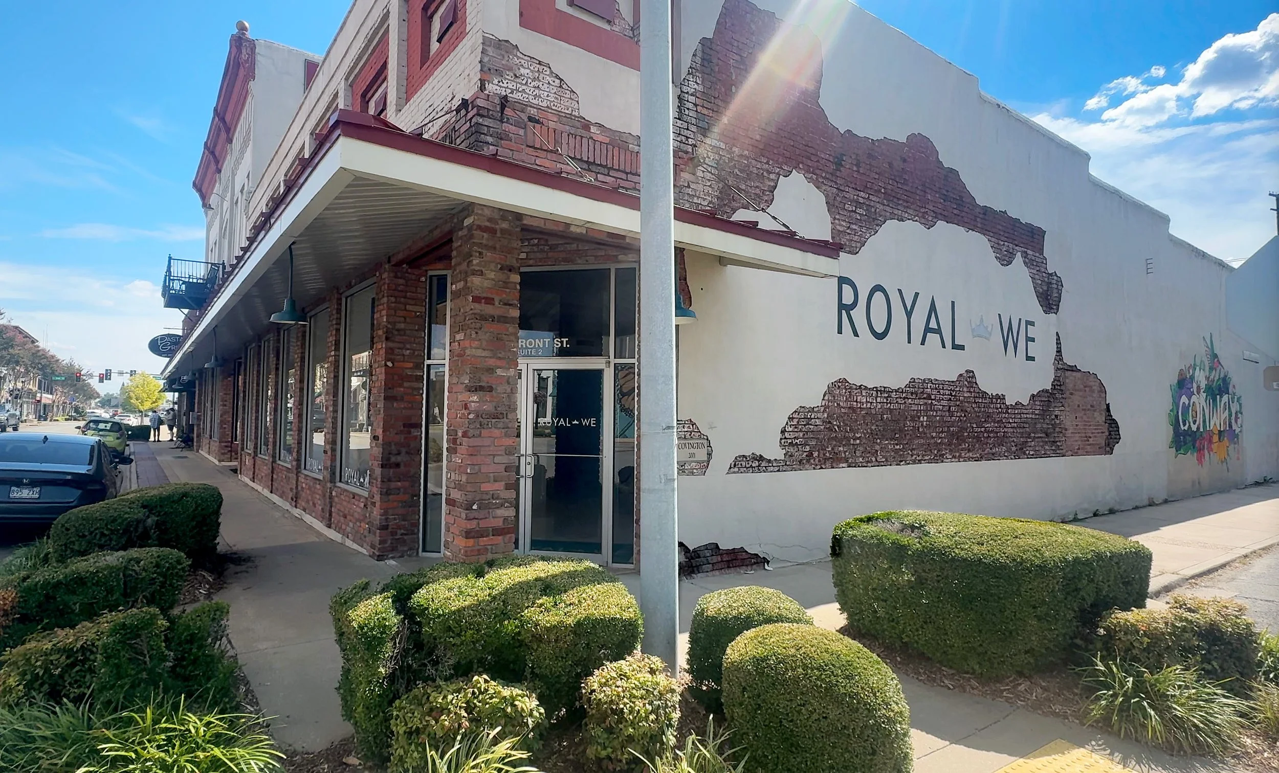 Street view of a brick building with a mural on the side. The mural depicts a map outline with the text "ROYAL WE" and a small crown. Bushes are in front of the building, and parked cars are along the street.