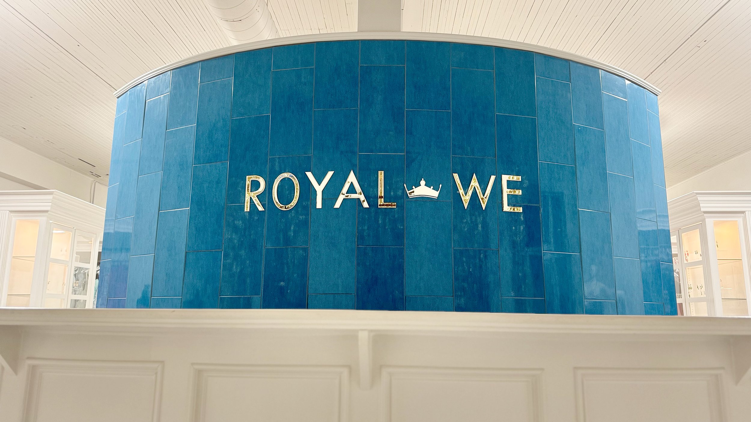 Interior of a store or mall with a large blue cylindrical display wall. The wall features the word 'ROYAL' in large silver letters, a crown logo between the words, and the word 'WE' on the right, also in silver letters.