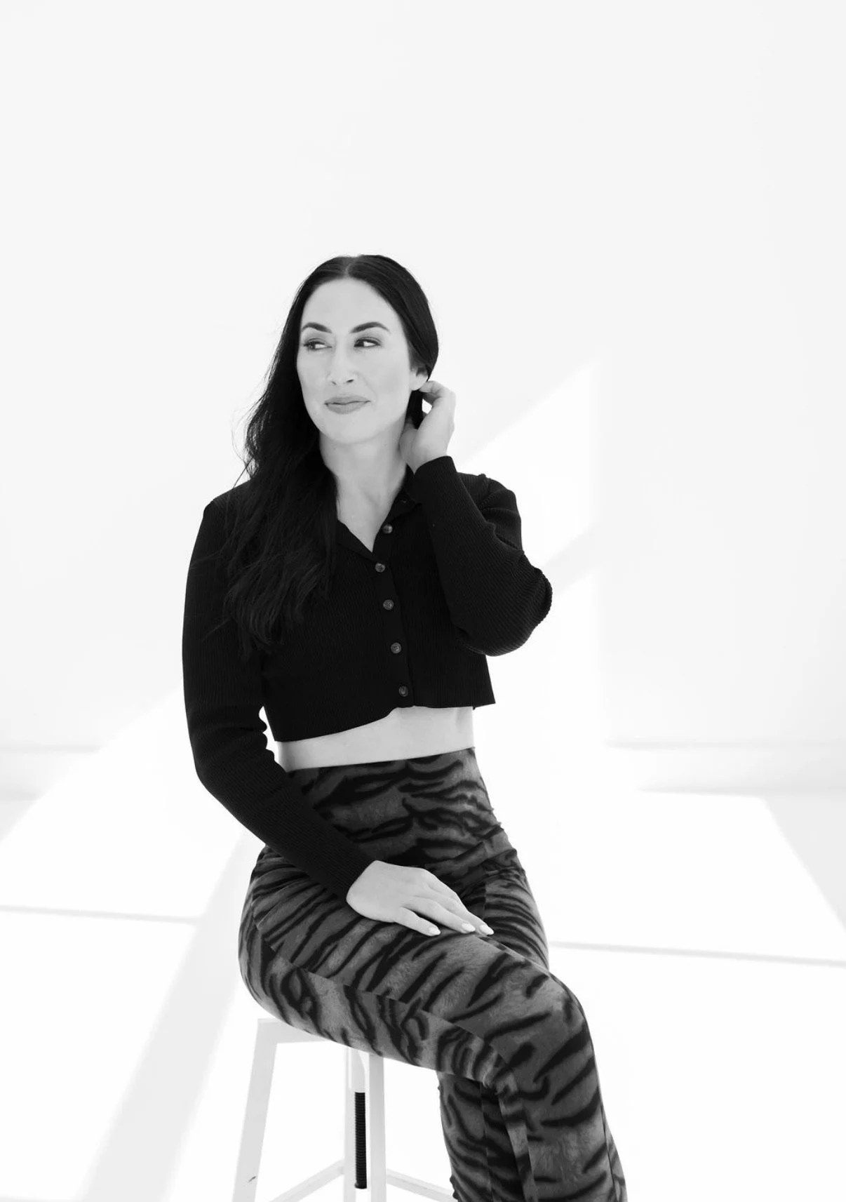 A woman with long dark hair, wearing a black cropped button-up sweater and tiger-striped pants, sitting on a white stool against a minimalistic white background.