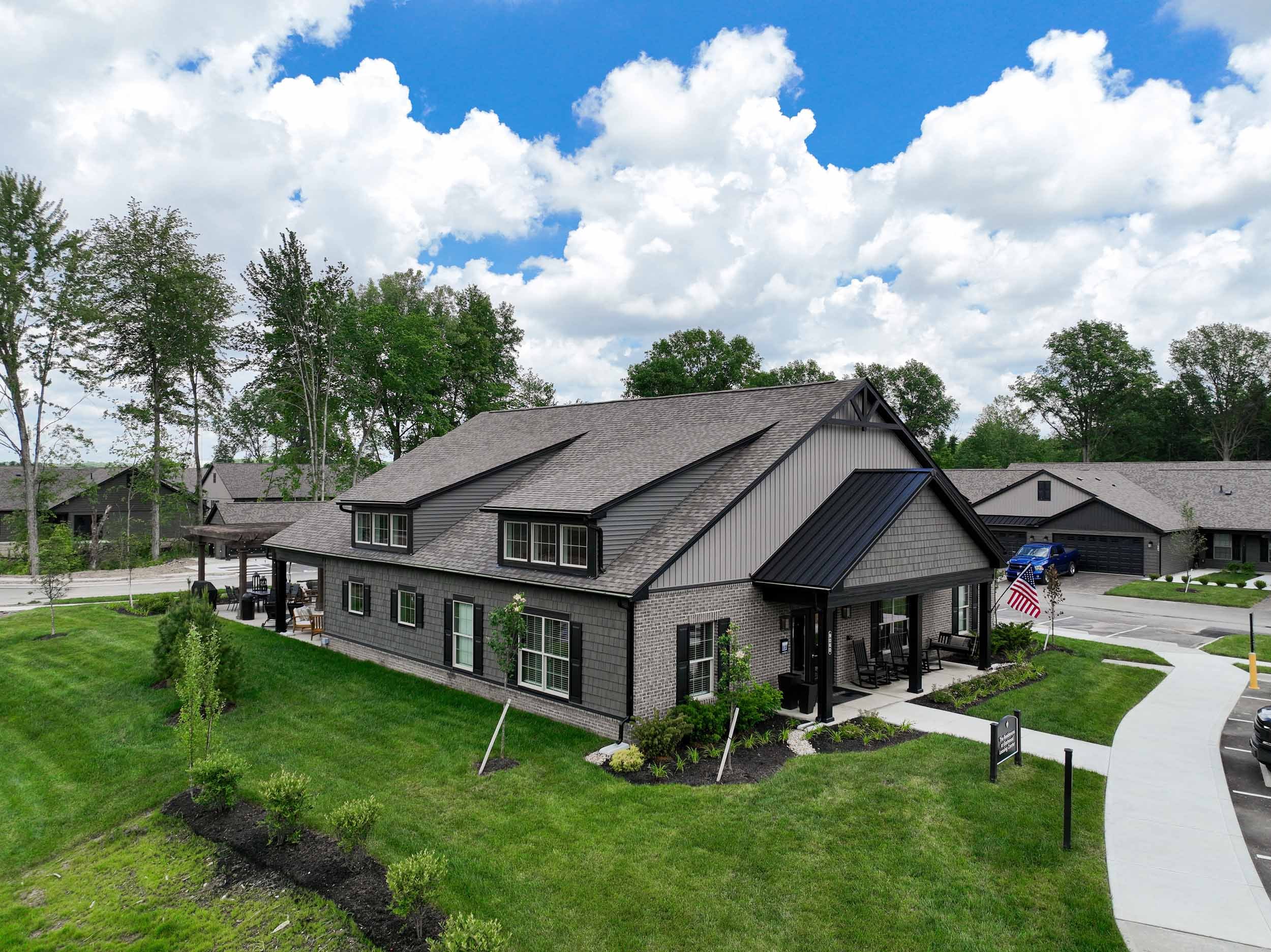 Modern residential house with gray exterior, black accents, a front porch with seating, surrounded by a well-manicured lawn, and trees in the background under a partly cloudy sky.