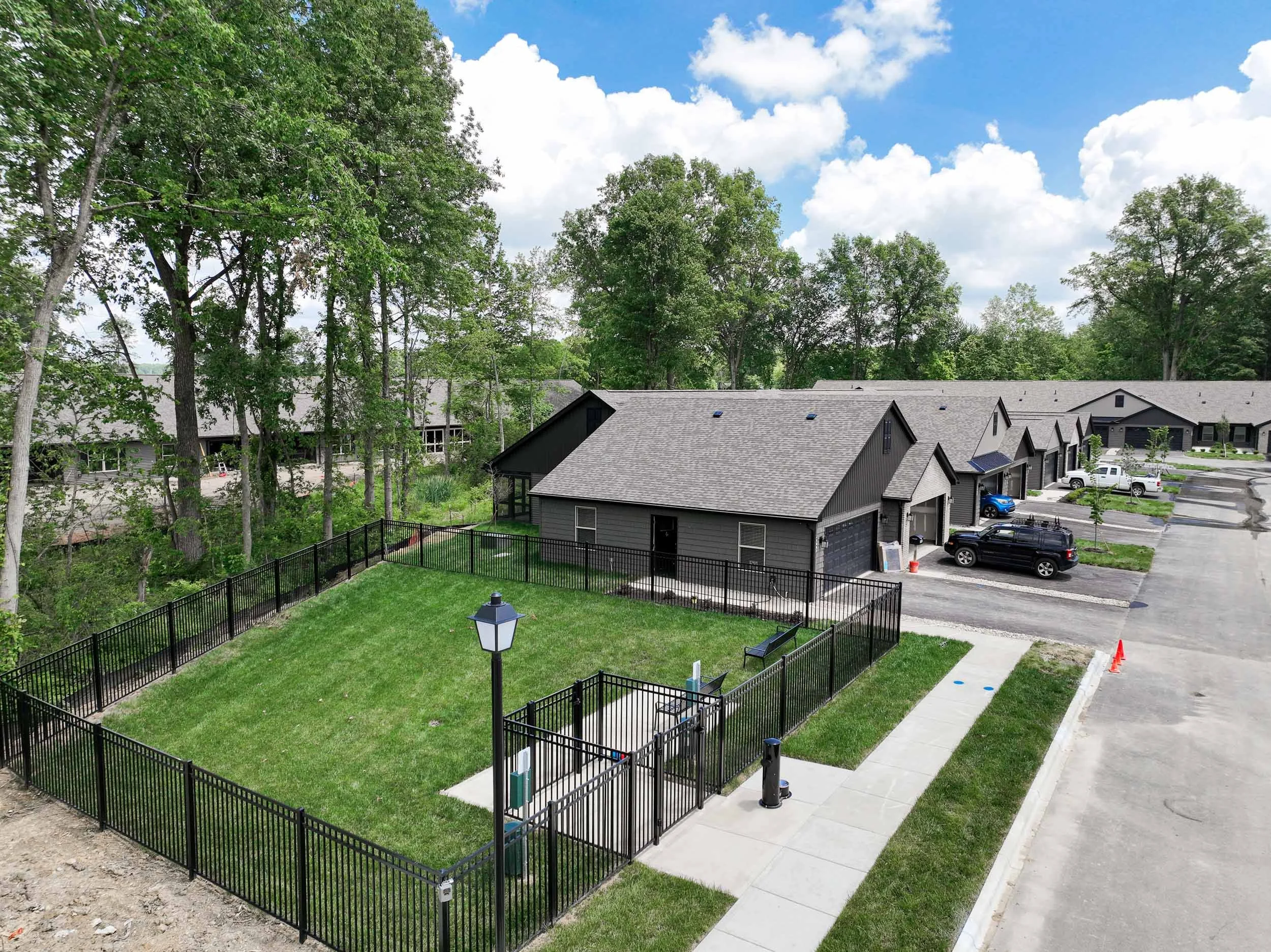 A fenced grassy yard with a picnic table, a lamp post, and a bench, located behind a row of townhouses with parking lots and cars, surrounded by trees under a partly cloudy sky.