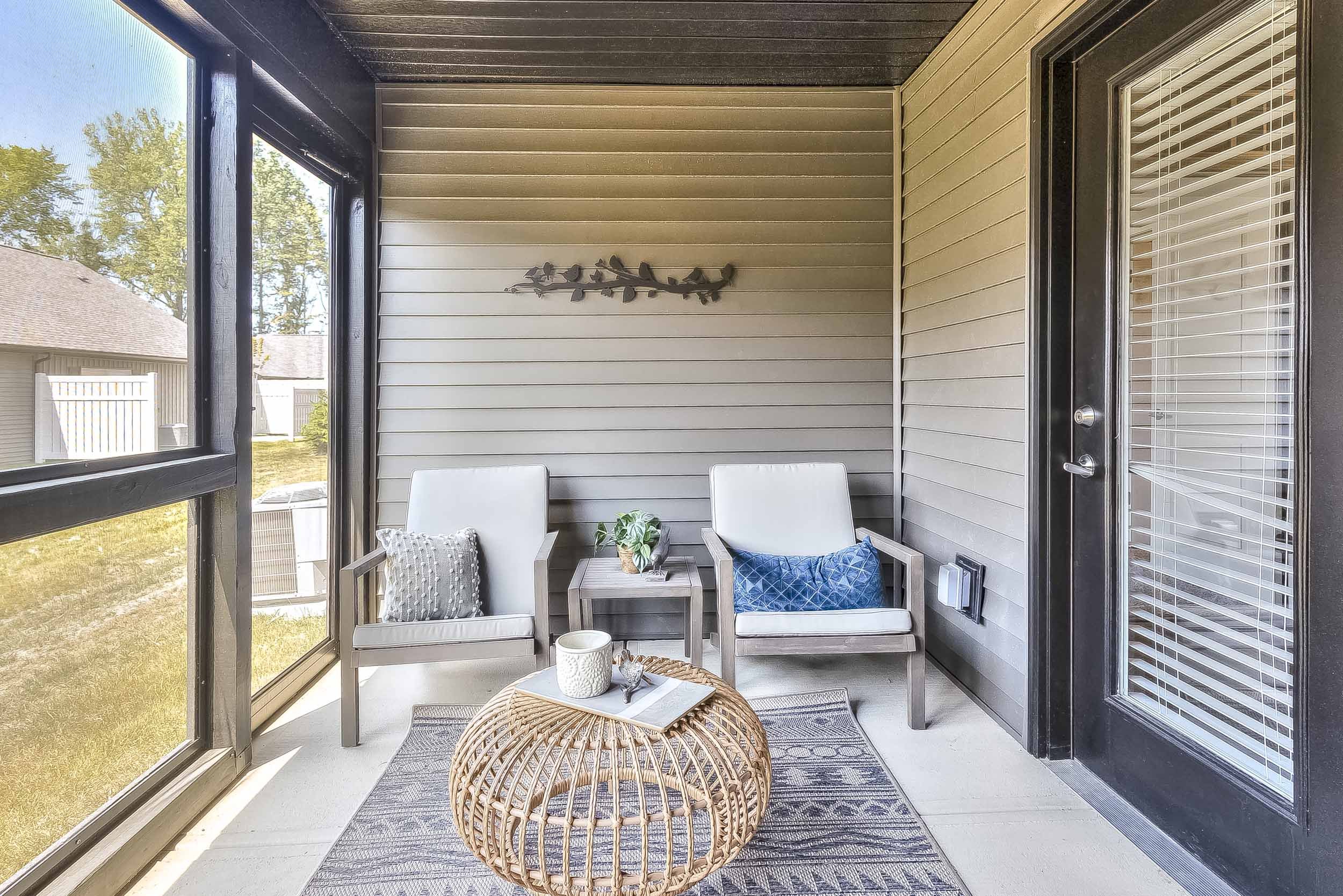 A cozy screened porch with two white armchairs, a small side table, decorative pillows, a woven coffee table, a patterned rug, and a wall decoration of birds on a wire. There is natural light coming in from the open screened sides.
