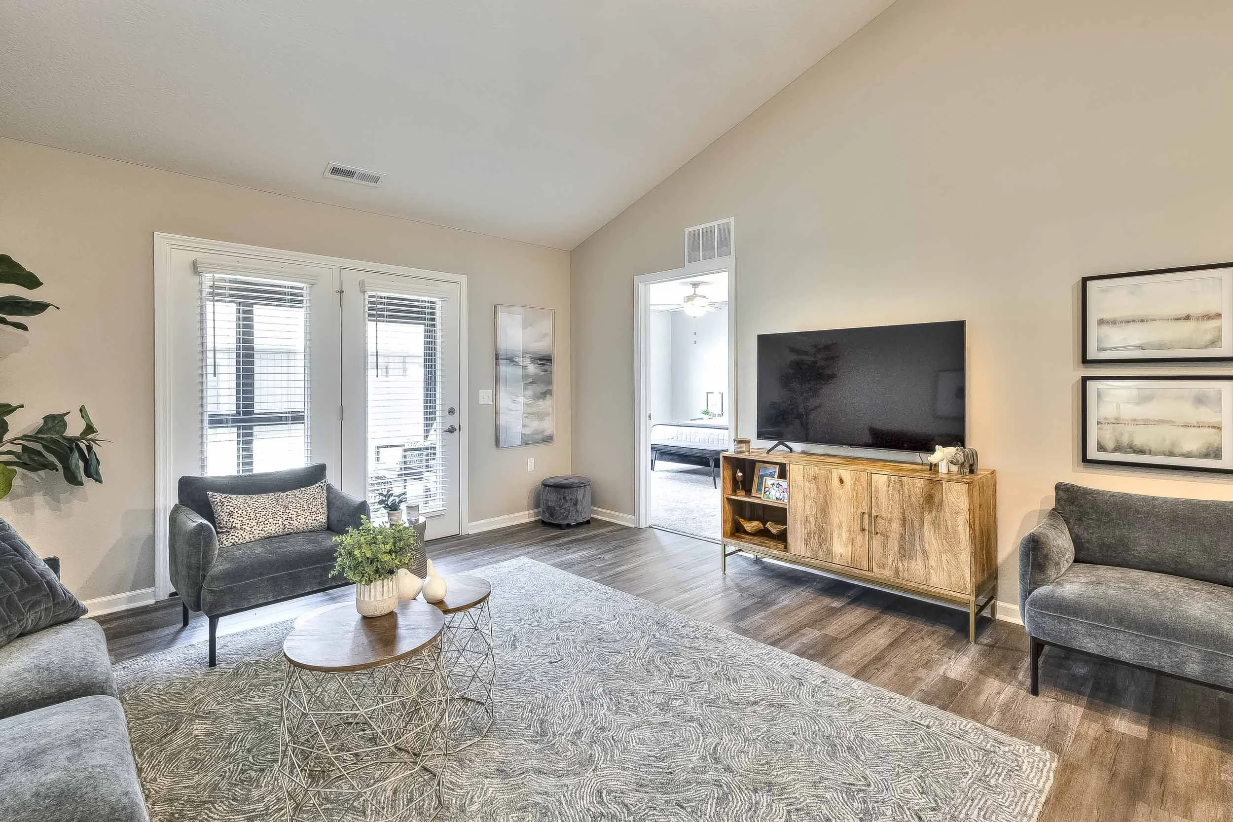 Living room with beige walls, gray furniture, wooden TV stand, large flat-screen TV, area rug, framed artwork, glass doors with blinds, and a view into a bright room with a ceiling fan.