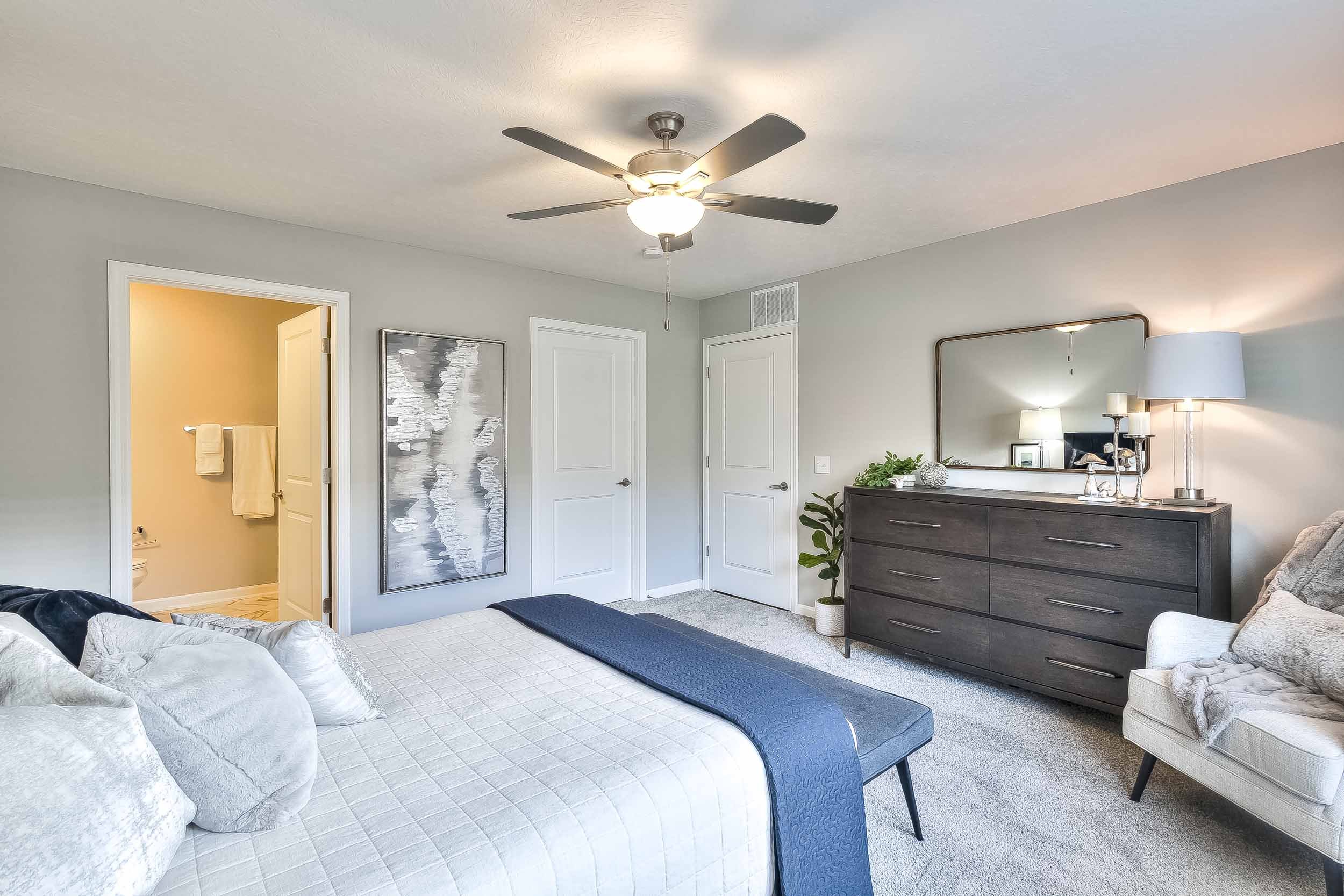 A bedroom with a bed, dresser with mirror, lamp, and chair, with an open door leading to a bathroom.