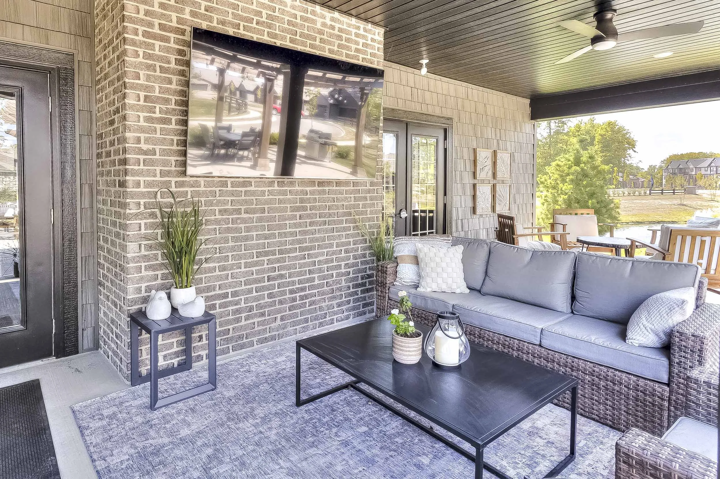 Outdoor patio with a gray wicker sofa, black coffee table, potted plants, a large television, and decorative wall art, overlooking a green yard and houses in the distance.