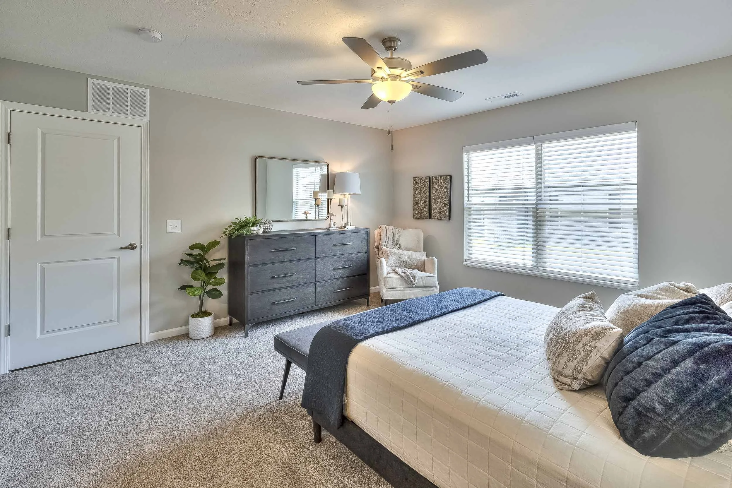 A bedroom with a bed, dresser, armchair, and window blinds, decorated in neutral colors.