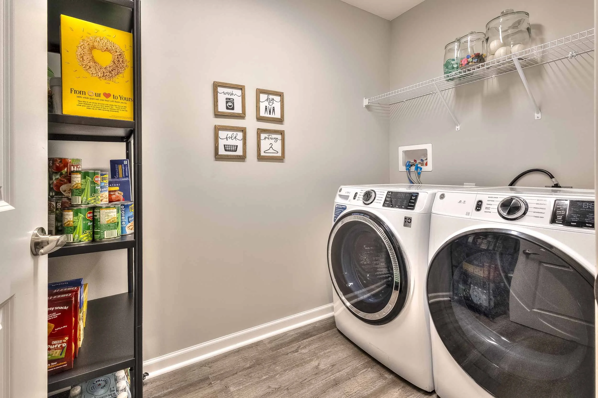 A laundry room with a washer and dryer side by side. A wire shelf above holds glass jars filled with various items. On the left wall, four framed prints with laundry-themed illustrations hang. To the left, a black shelving unit contains canned goods 