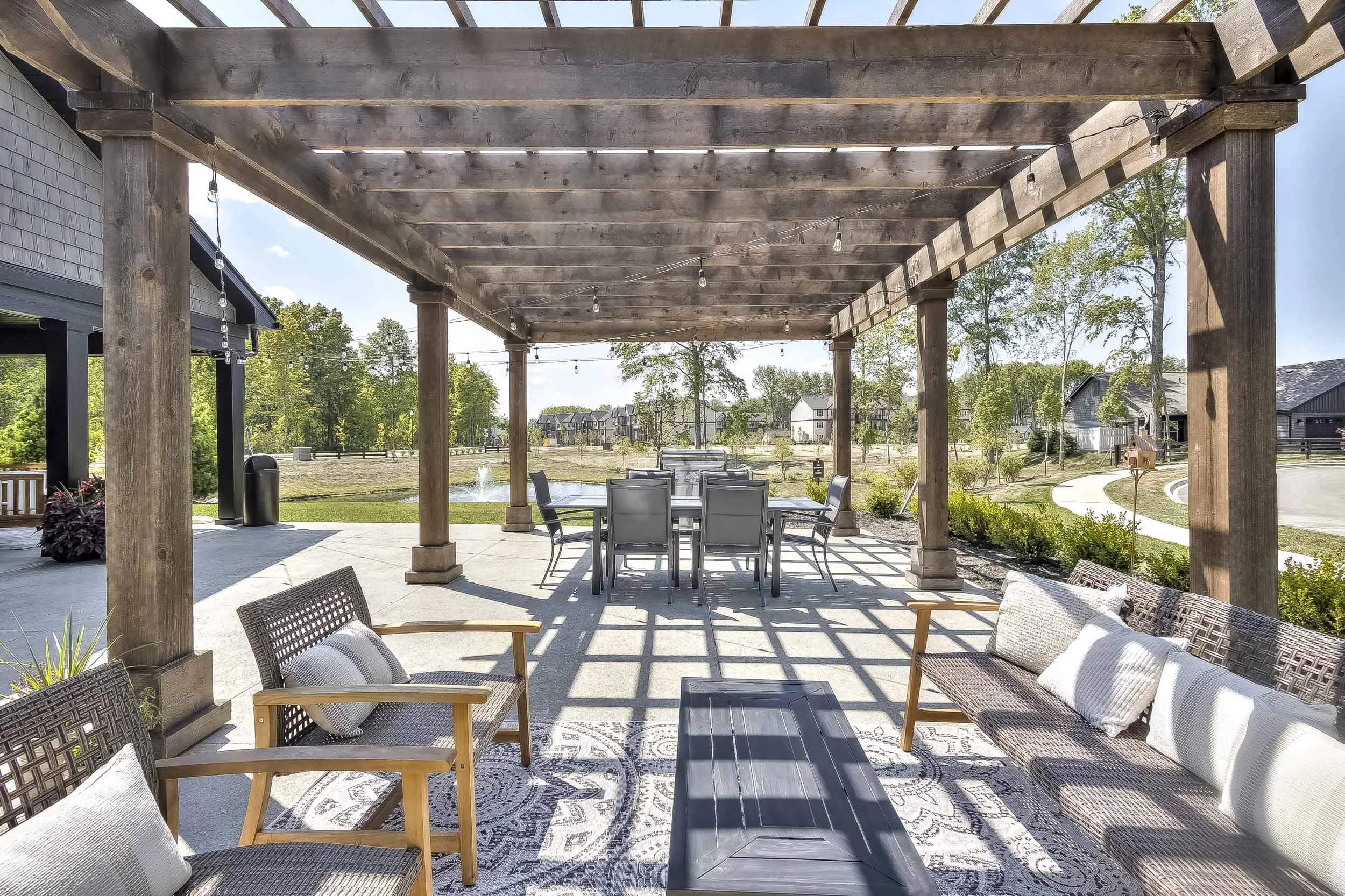 Outdoor patio with wooden pergola, seating area with cushioned chairs, and a dining table, overlooking a landscaped yard with trees, a pond with fountain, and neighboring houses in the background.