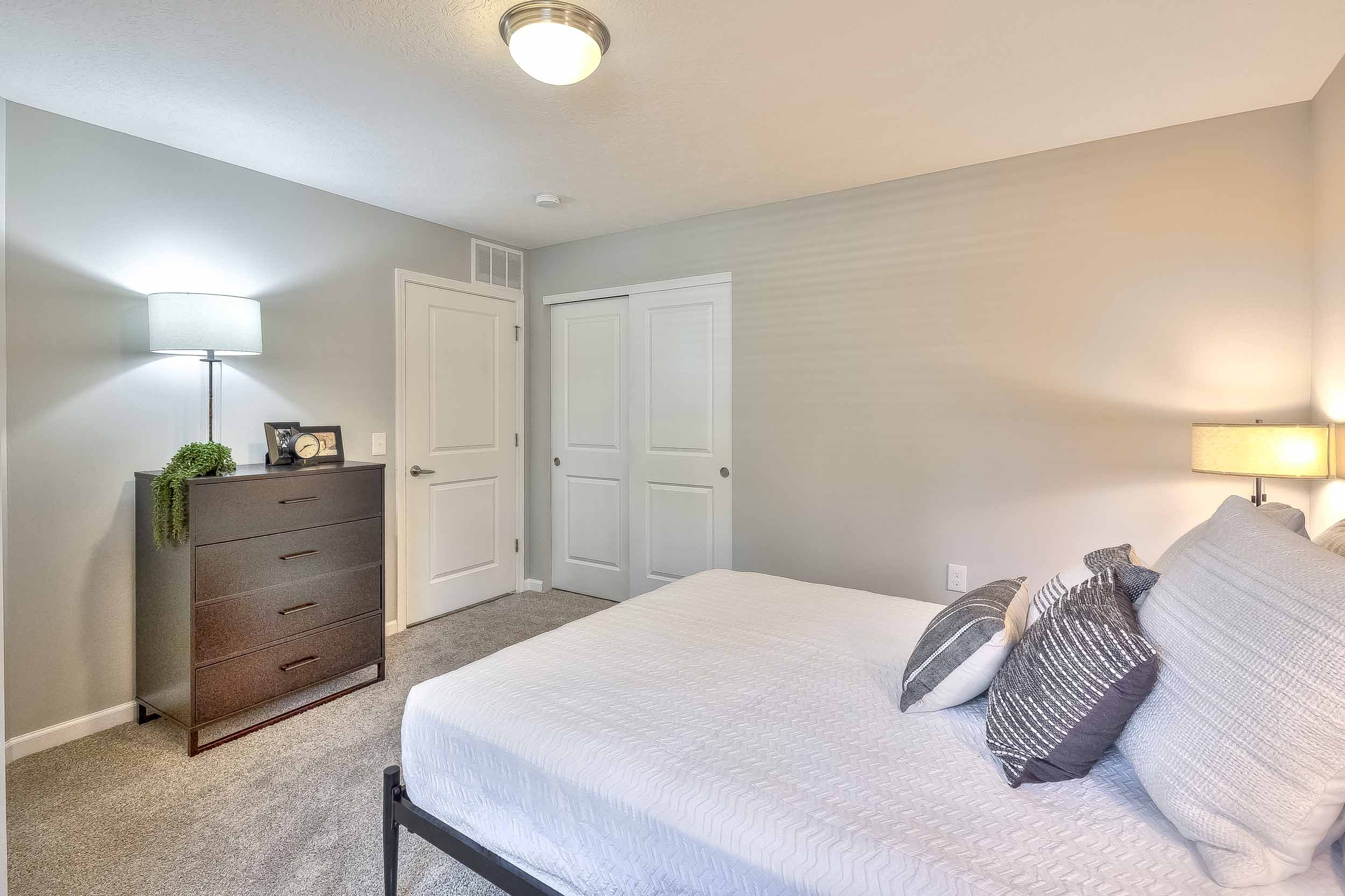 A bedroom with a bed, nightstand, dresser, and closet doors, illuminated by bedside and overhead lights.