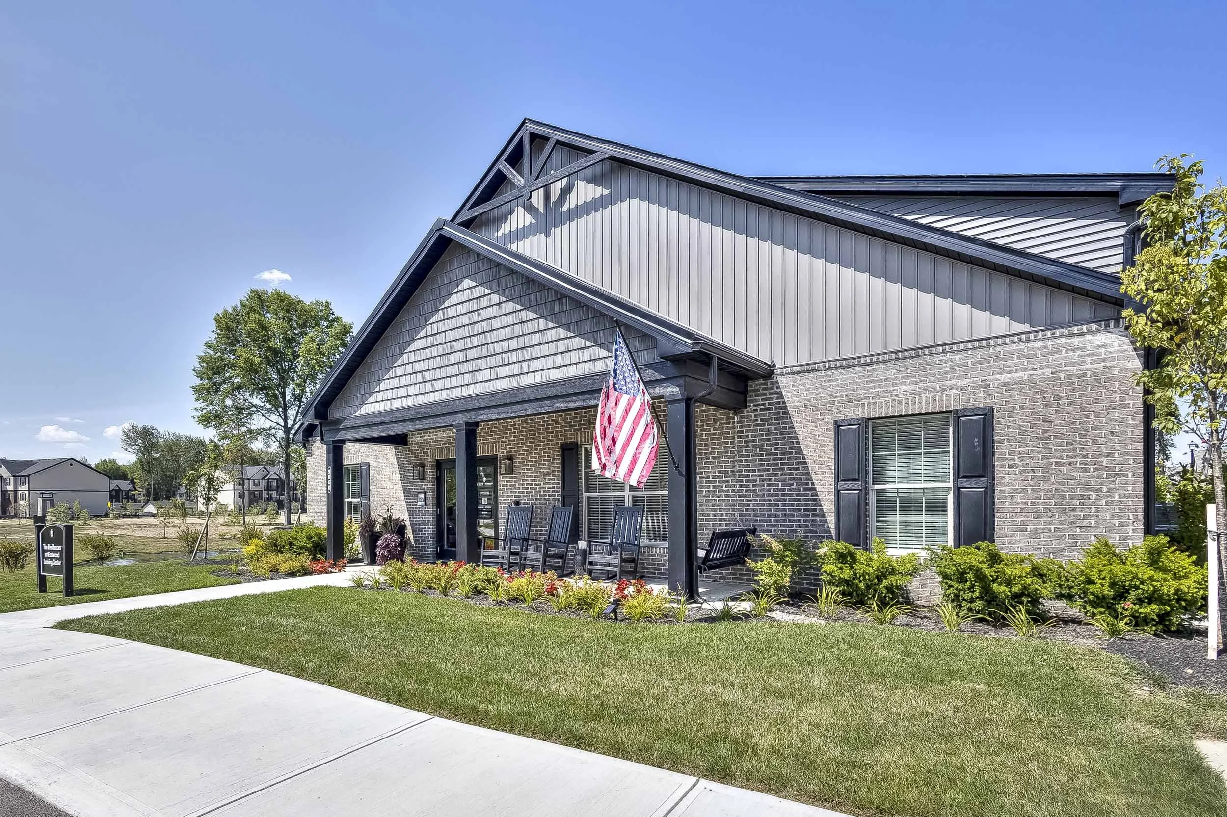A modern brick and gray siding residential building with a covered porch, black rocking chairs, and an American flag, surrounded by a well-maintained lawn and shrubs.