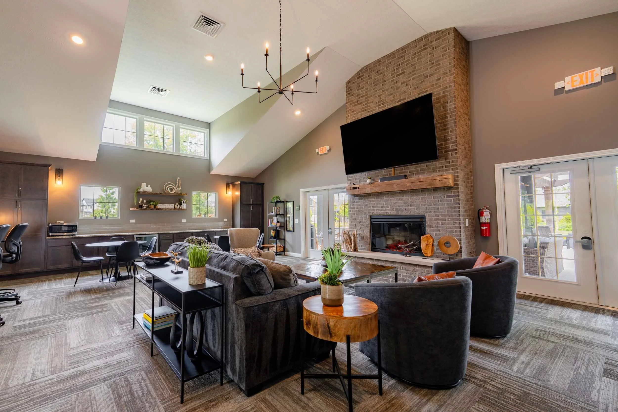 Spacious living area with high vaulted ceiling, brick fireplace with a large TV above, and several windows letting in natural light. Furnished with sofas, a coffee table, plants, and a mix of modern and rustic decor.