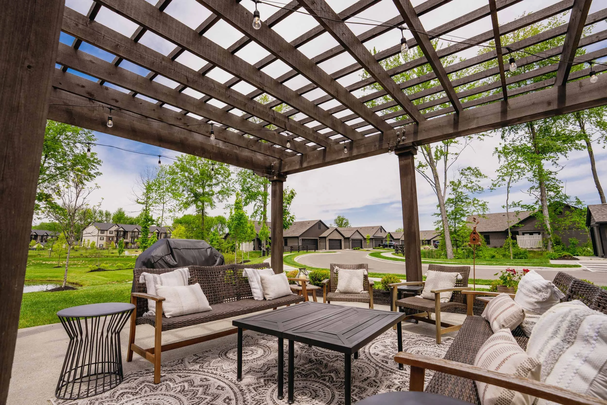 Patio with outdoor furniture including wicker sofas and chairs with cushions, a black coffee table, and a patterned outdoor rug, all shaded by a wooden pergola with string lights, overlooking a suburban neighborhood with trees, houses, and a street.