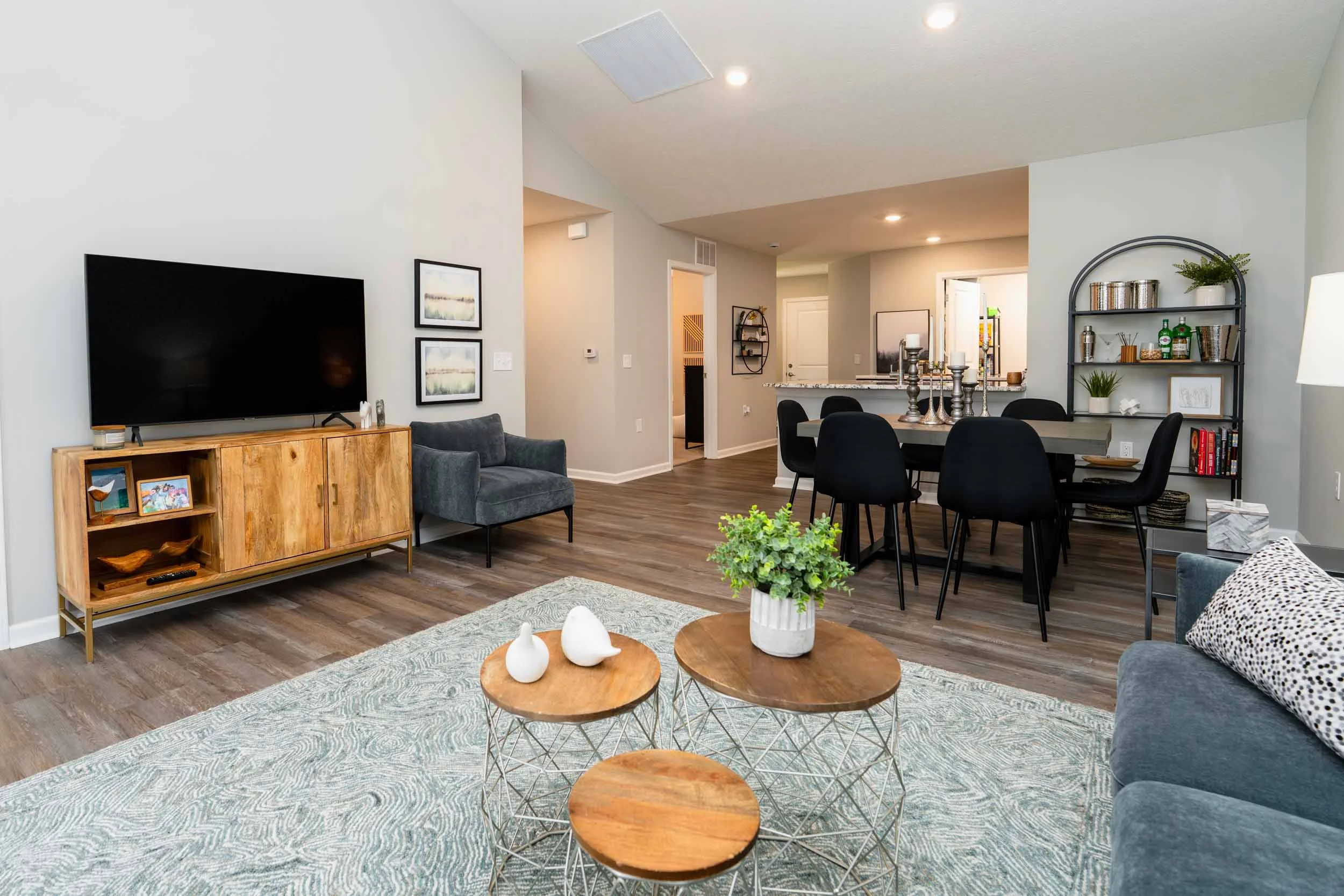 Living room with a wooden TV stand, a flat-screen TV, grey armchair, framed art, black dining table with six chairs, black shelf with decor, potted plants, candles, and a patterned pillow, hardwood flooring, and neutral-colored walls.